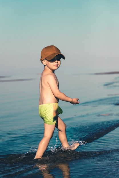 Een jongen in een groene korte broek en een bruine baseballpet loopt op de zee in de buurt van de kust Kopieer de ruimte