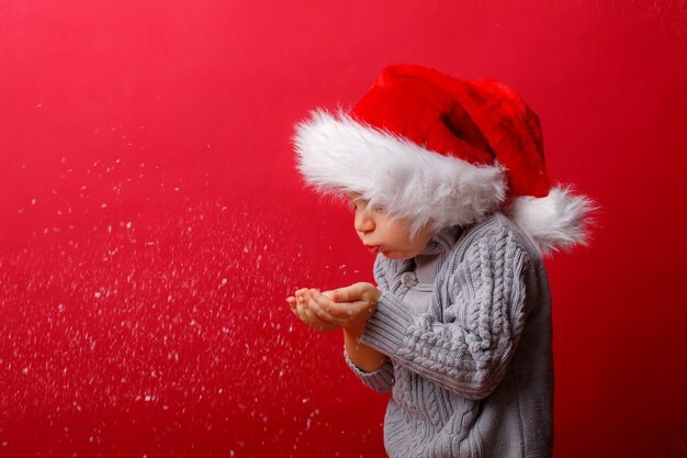 Een jongen in een kerstman hoed blaast sneeuw op een rode achtergrond