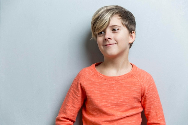 Een jongen in een oranje shirt zit tegen een witte muur.