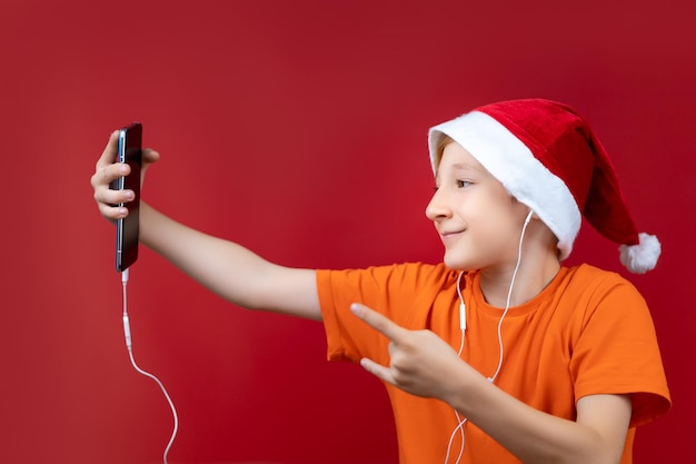 Een jongen in een oranje T-shirt en een rode kerstmuts houdt een telefoon voor zich en gebaart ernaar