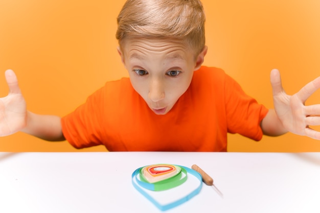 Een jongen in een oranje T-shirt zit aan een witte tafel en heft emotioneel zijn handen op en kijkt naar het werk dat is gedaan met dunne papieren stroken in de quilling-techniek