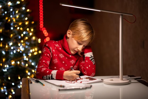 Een jongen in een rode kersttrui op de achtergrond van een kerstboom drukte zijn hand tegen zijn gezicht en tekende bedachtzaam