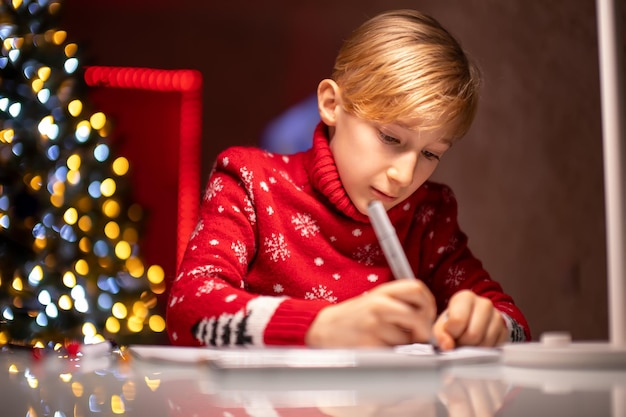Een jongen in een rode kersttrui op de achtergrond van een kerstboom is enthousiast bezig met tekenen