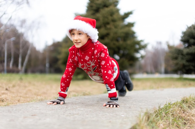 Een jongen in een warme kersttrui met een nieuwjaarshert en een kerstmuts maakte zich klaar om een push-upoefening uit te voeren in een stadspark op de sporen