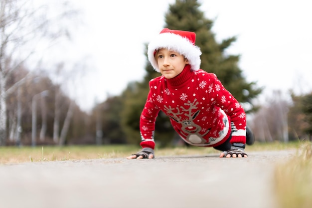 Een jongen in een warme kersttrui met een nieuwjaarshert en een kerstmuts traint door opdrukoefeningen te doen in een stadspark op de baan