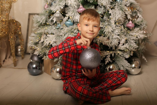 een jongen in nieuwjaarspyjama zit bij de kerstboom met een nieuwjaarsspeeltje in zijn handen