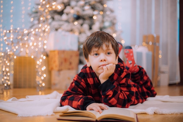Een jongen in pyjama leest een boek onder een kerstboom. De stemming van het nieuwe jaar. Boeken lezen . Kinderboeken.