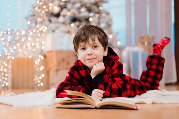 Een jongen in pyjama leest een boek onder een kerstboom. Nieuwjaarsstemming. Boeken lezen . Kinderboeken. Een cadeau voor het nieuwe jaar