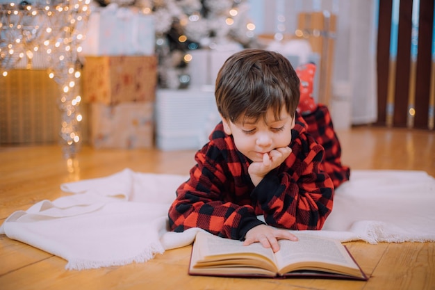 Een jongen in pyjama leest een boek onder een kerstboom. Nieuwjaarsstemming. Boeken lezen . Kinderboeken.