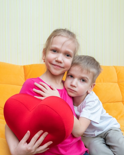 Een jongen knuffelt zachtjes een meisje met een rood hartvormig kussen thuis. Familie feest. Gefeliciteerd met je verjaardag, fijne Valentijnsdag, fijne familiedag