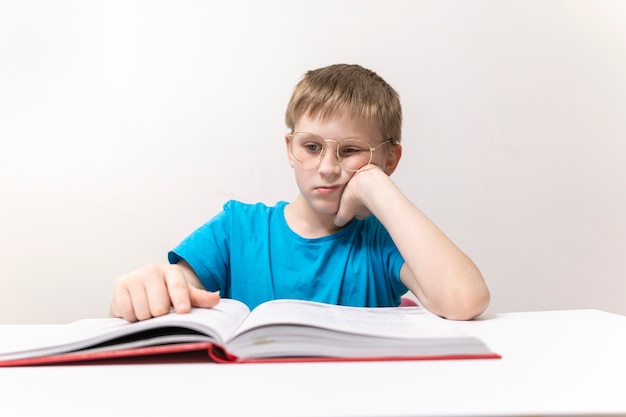 Een jongen leest een boek a met een bril op een witte tafel en een witte muurachtergrond