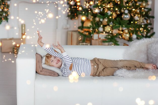 Een jongen met blond haar tegen de achtergrond van een kerstboom ligt op een bank en droomt van het beste kerstcadeau