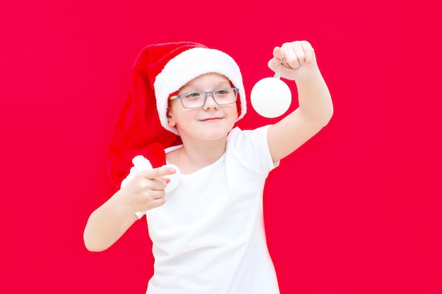 Een jongen met een kerstmuts houdt een kerstbal in zijn handen, wijst ernaar en glimlacht