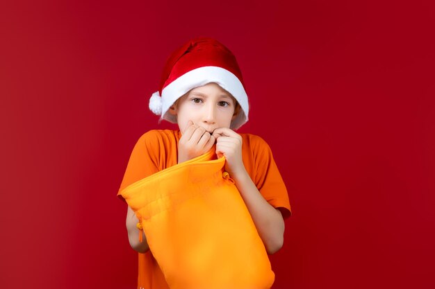 Een jongen met een kerstmuts op een rode kerstachtergrond houdt een gele cadeautas in zijn handen en kijkt angstig in de camera