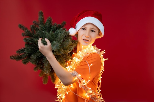 Een jongen met een rode kerstmuts en een kerstslinger om zijn nek die gloeit, draagt een kerstboom voor de feestdagen
