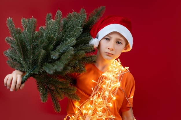 Een jongen met een rode kerstmuts en een kerstslinger om zijn nek houdt een kerstboom op zijn schouder