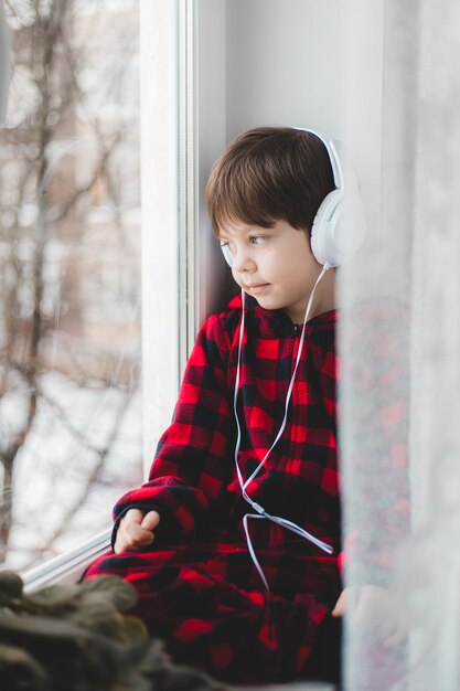 Een jongen met koptelefoon kijkt uit het raam. Moderne kinderen. Sociale media. Een artikel over moderne kinderen.
