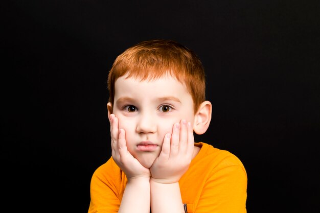 Een jongen met rood haar houdt zijn handen voor zijn gezicht, een jongen met een mooi gezicht tegen een donker