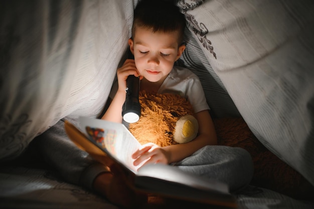 Een jongen van 56 jaar leest 's avonds in het donker een boek onder een deken met een speelgoedbeer