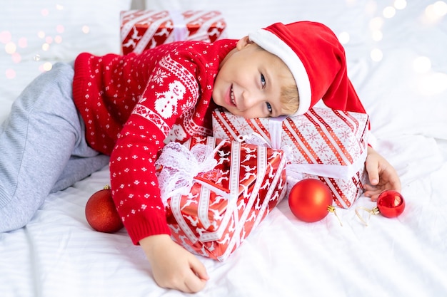 Een jongenskind met een kerstmuts en een rode trui ligt met nieuwjaarscadeaus op een bed nieuwjaarskerst