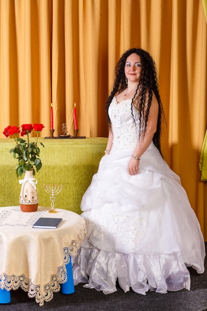 Een joodse bruid in een witte trouwjurk zonder sluier staat aan een tafel met bloemen voor de choepa-ceremonie