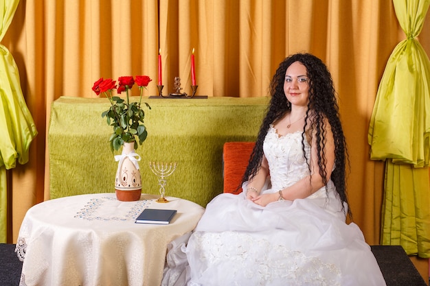 Een Joodse bruid in een witte trouwjurk zonder sluier zit aan een tafel met bloemen voor de choepa-ceremonie. Horizontale foto