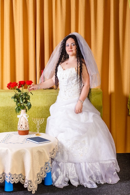 Een Joodse bruid met zwarte krullen in een witte jurk met een sluier staat in de kamer te wachten op de bruidegom voor de choepa-ceremonie