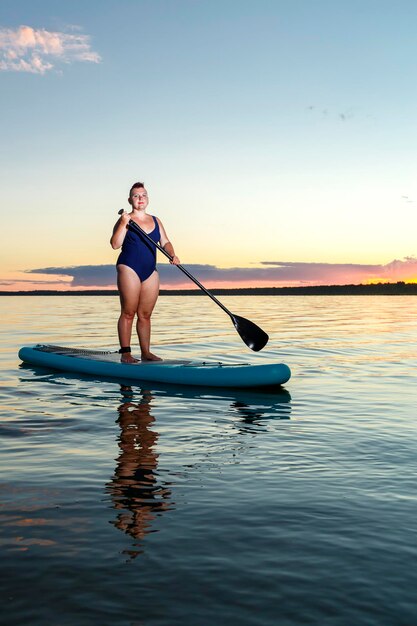 Een joodse feministische vrouw in een gesloten zwempak met een mohawk die op een SUP-bord staat met een roeispaan drijft op het water tegen de achtergrond van de avondrood