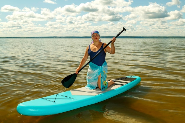 Een Joodse vrouw in een gesloten zwempak een pareo en een hoofdtooi op haar knieën op een SUP board met een peddel op het meer