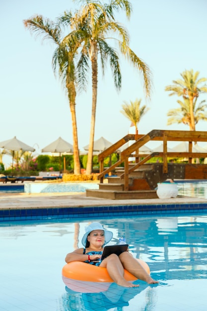 Een joodse vrouw met een zonnehoed in het zwembad in een zwemkring met een cocktail en een laptop op een achtergrond van palmbomen.