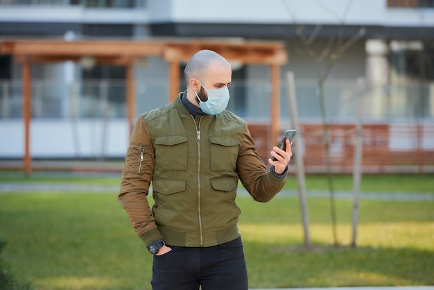 Een kale man in een medisch gezichtsmasker om te voorkomen dat het verspreide coronavirus zijn smartphone controleert in de gezellige straat.