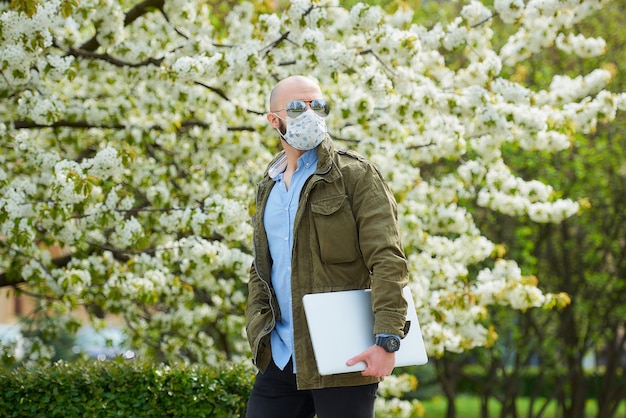 Een kale man met een baard in een medisch gezichtsmasker om de verspreiding van het coronavirus te vermijden, loopt met een laptop in het park. Een man draagt een gezichtsmasker n95 en een pilotenzonnebril in de tuin tussen bloeiende bomen.