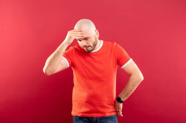 Een kale man van middelbare leeftijd met een baard en in een rood T-shirt. Houdt zijn hoofd vast met zijn handen. Hij heeft hoofdpijn.