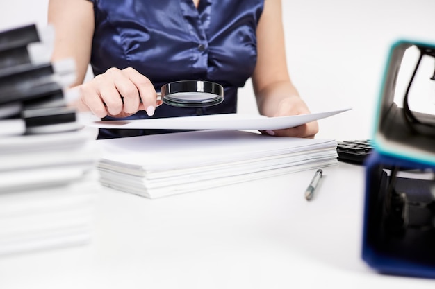 Een kantoormedewerker onderzoekt documenten door een vergrootglas op de achtergrond van stapels documenten