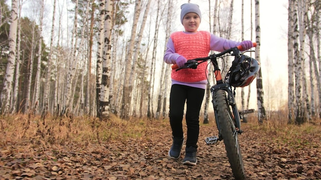 Een kaukasische kinderen lopen met de fiets in het herfstpark Klein meisje lopen zwart oranje fiets in het bos Kid gaat fietssporten Biker motion ride met rugzak en helm Mountainbike hardtail