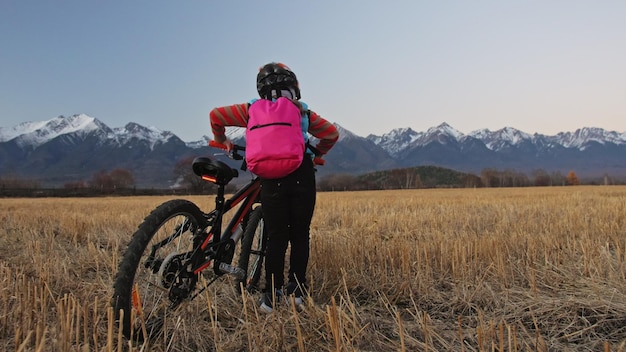 Een kaukasische kinderen lopen met fiets in tarweveld meisje lopen zwart oranje cyclus op de achtergrond van prachtige besneeuwde bergen biker motion rit met rugzak en helm mountainbike harde staart