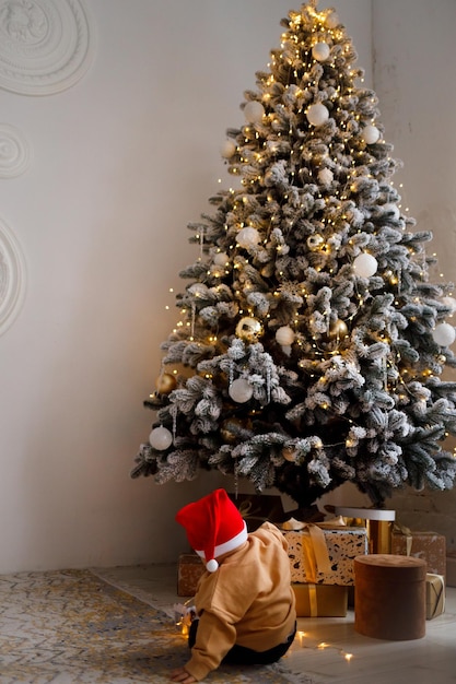 Een kerstjongen met een kerstmuts bij een versierde kerstboom speelt met zijn cadeautjes