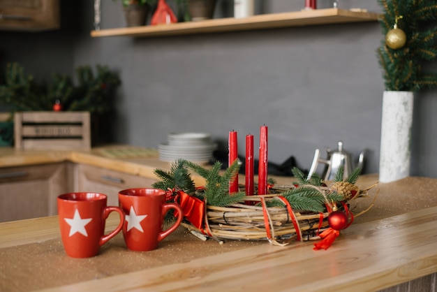 Een kerstkrans met rode kaarsen op de houten tafel en twee rode mokken in de keuken,