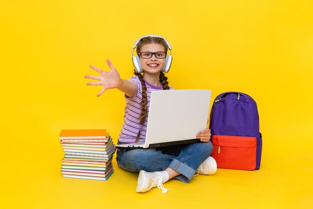 Foto een kind dat koptelefoons draagt met een laptop toont zijn handpalm die naar voren strekt een jong meisje met boeken en een rugzak doet online huiswerk met een leraar gele geïsoleerde achtergrond