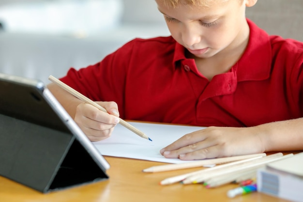Een kind dat voor een tablet zit met papier en kleurpotloden tekent of voltooit een taak op internet thuisonderwijs online soft focus