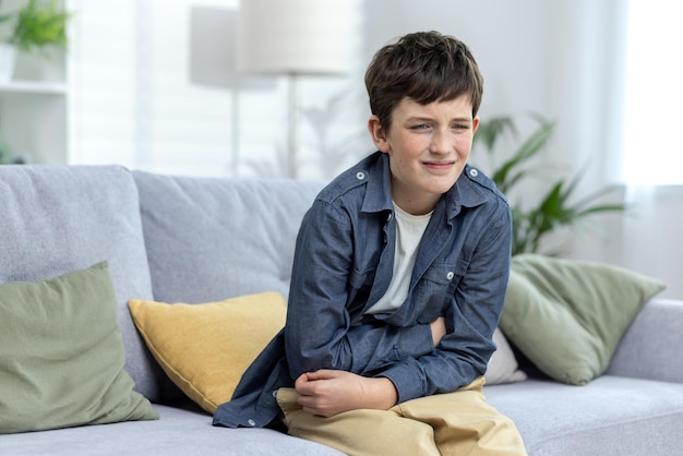 Een kind een jongen een tiener zit alleen thuis op de bank en houdt zijn buik vast met zijn handen