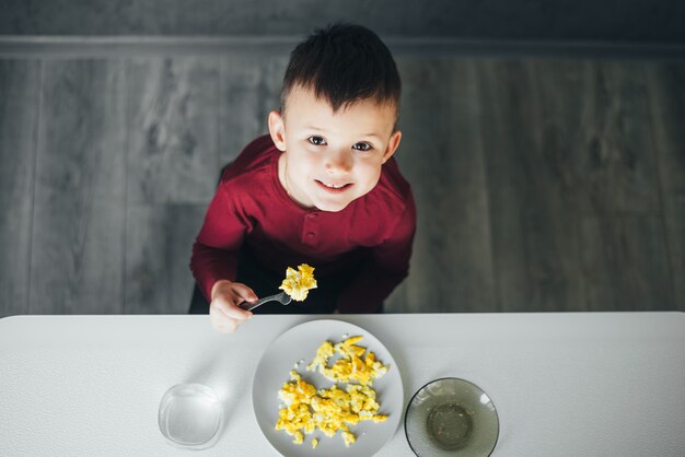 Een kind in de namiddag op een wit-lichte keuken in een bordeauxrode trui eet een omelet