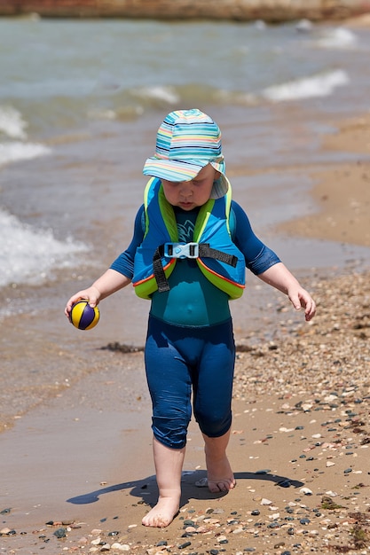 Een kind in een zwemvest en een bal in de hand loopt langs het water langs het zeestrand