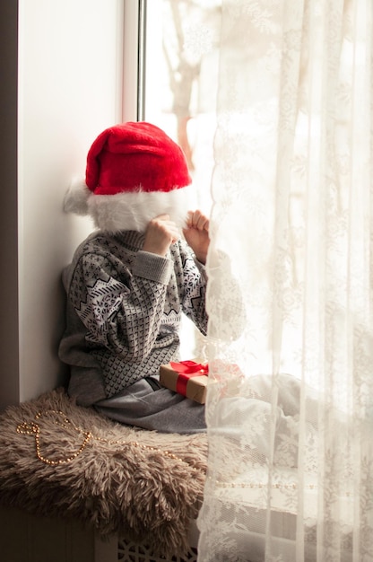 Een kind in feestelijke kleding en een rode kerstmuts zit bij het raam en bedekt zijn gezicht met een hoed