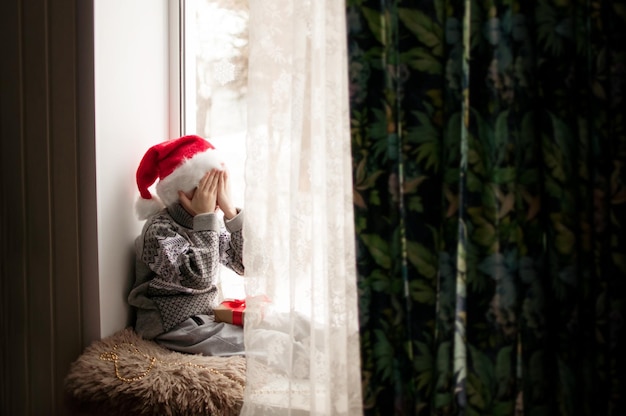 Een kind in feestelijke kleding en een rode kerstmuts zit bij het raam en bedekt zijn gezicht met zijn handen