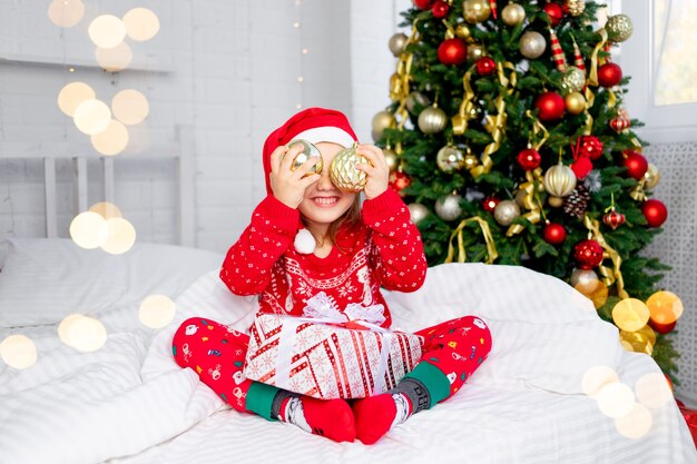 Een kind meisje met kerstballen op haar oren bij de kerstboom in een rode trui en kerstman hoed op oudejaarsavond in een wit bed glimlachend, gek rond en lachen met een cadeau