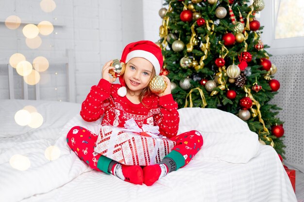 Een kind meisje met kerstballen op haar oren bij de kerstboom in een rode trui en kerstman hoed op oudejaarsavond in een wit bed glimlachend, gek rond en lachen met een cadeau