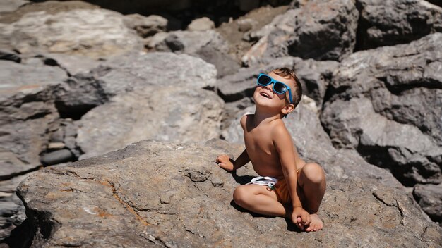 Een kind met een zonnebril zit en zonnebaadt op grote rotsblokken op het strand bij de zee