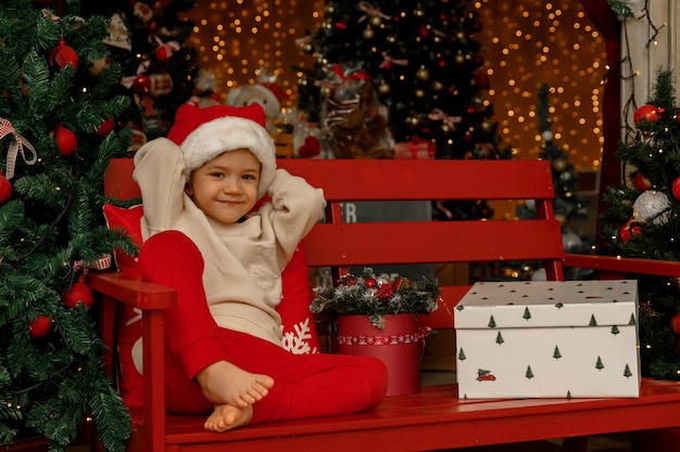 Een kind met kerstmuts zit op een bankje voor de kerstboom