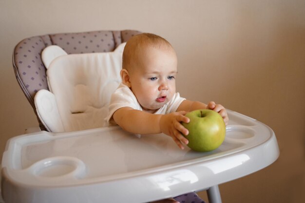 een kind op een kinderstoel in een wit rompertje speelt met een groene appel Eten